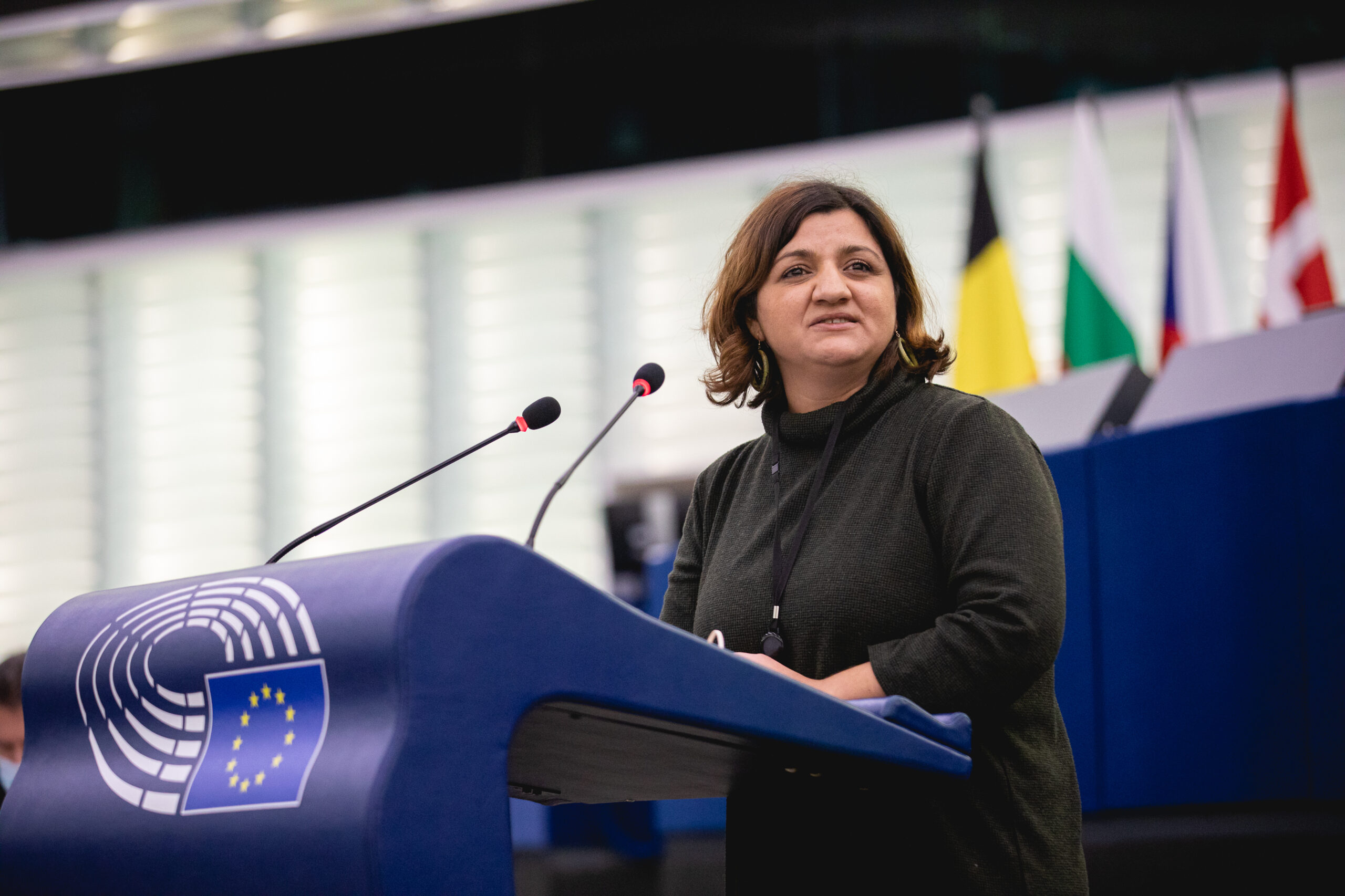 Özlem Demirel eine Rede im Plenum haltend. Blaues EU-Pult und schwarzer Anzug